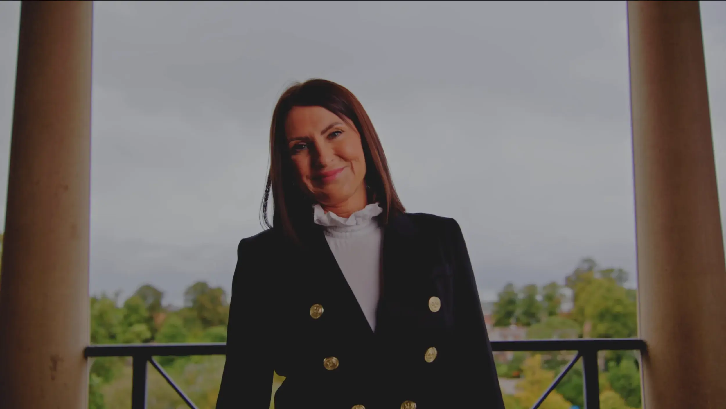 A woman in a black blazer with gold buttons stands on a balcony, overlooking a scenic landscape under cloudy skies.