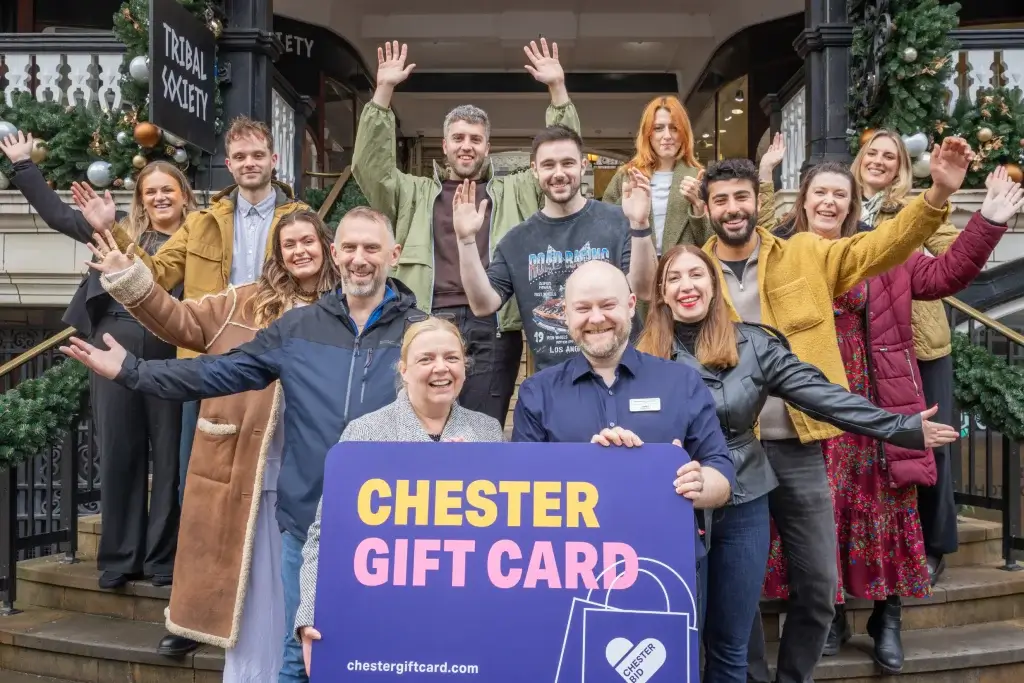 A group of diverse individuals stands on a staircase, celebrating with arms raised, holding a "Chester Gift Card" sign in front.