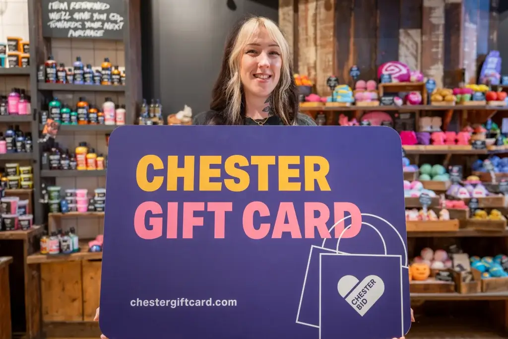 Smiling person holding a large purple sign reading CHESTER GIFT CARD in a shop with colourful bath products