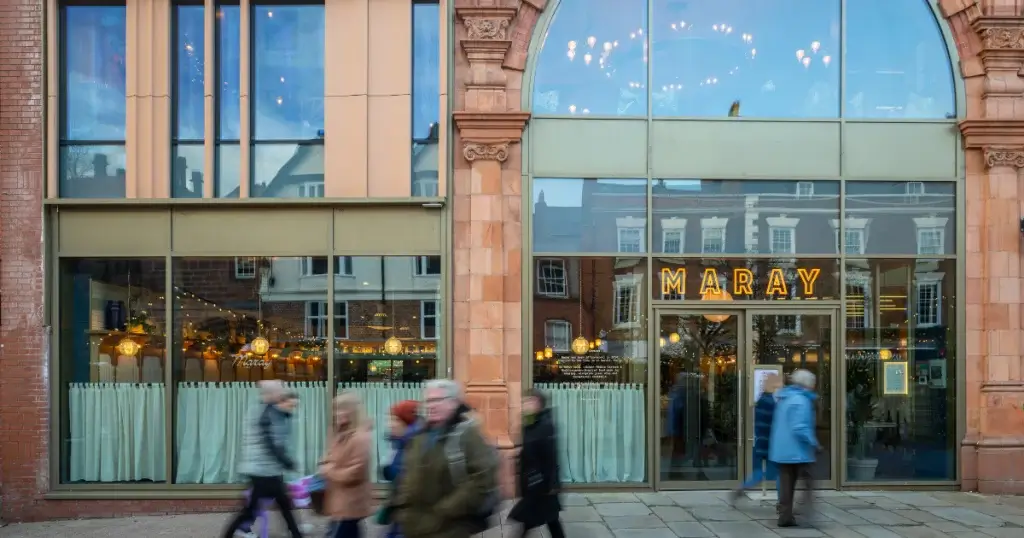 The exterior of Maray restaurant features large windows, decorative lighting, and onlookers walking by on a bustling street.