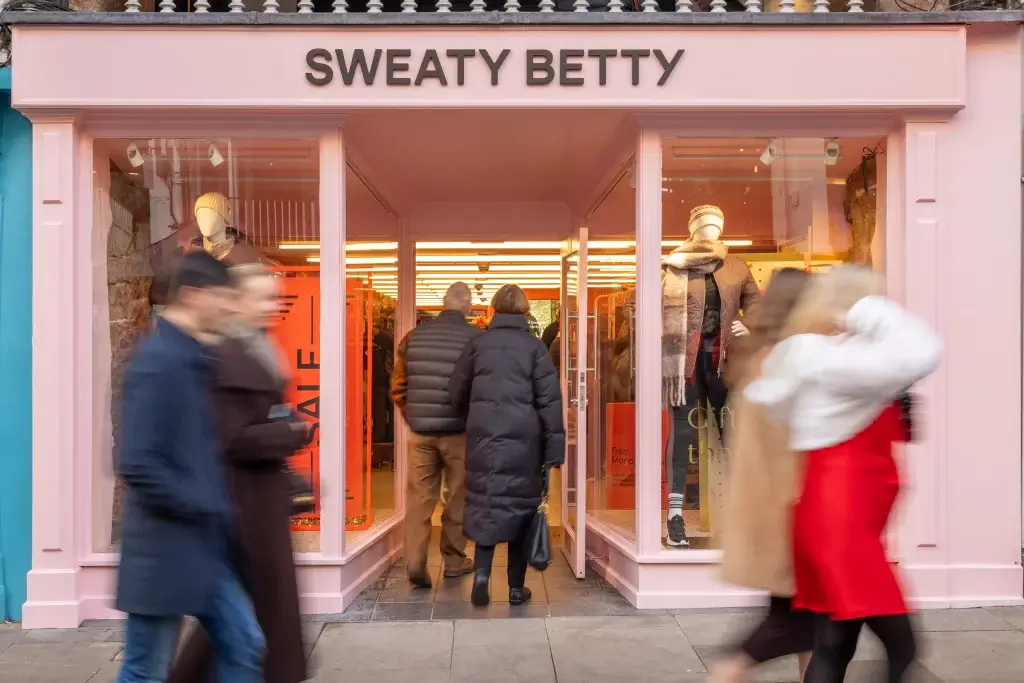 Entrance of a Sweaty Betty store with shoppers walking in, showcasing a pink facade and a mannequin dressed in winter apparel.