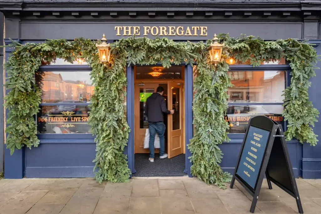 A man enters The Foregate pub, adorned with festive greenery and lanterns, welcoming guests during the holiday season.