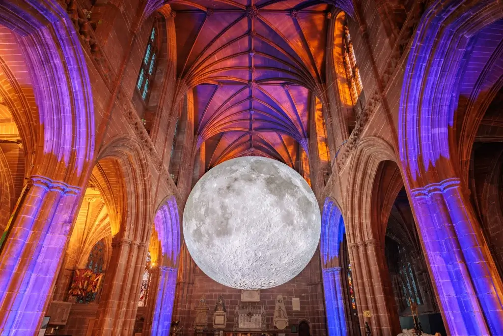 A large, realistic moon installation hangs inside a gothic cathedral, illuminated by colorful lights from the stained glass windows.