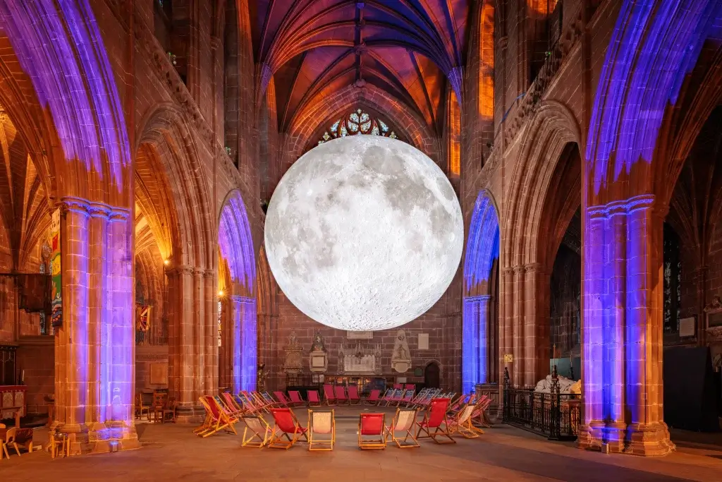 A large, realistic moon installation hangs inside a gothic cathedral, illuminated by colorful lights from the stained glass windows.