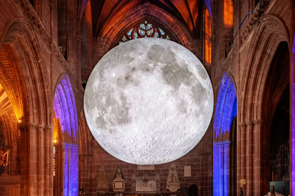 A large, realistic moon installation hangs inside a gothic cathedral, illuminated by colorful lights from the stained glass windows.