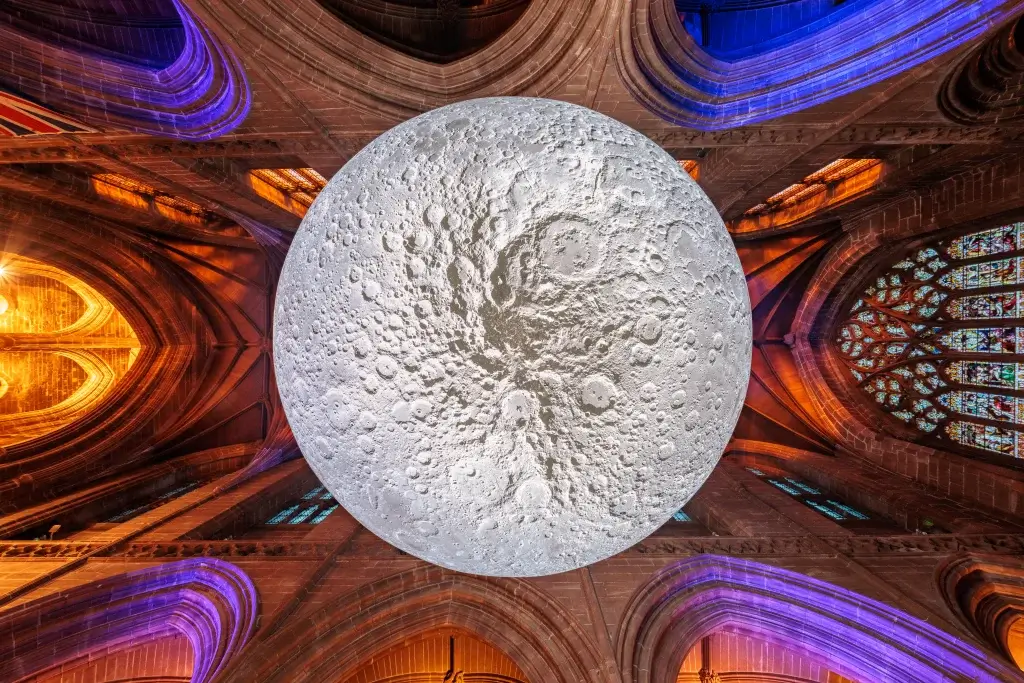 A large, realistic moon installation hangs inside a gothic cathedral, illuminated by colorful lights from the stained glass windows.