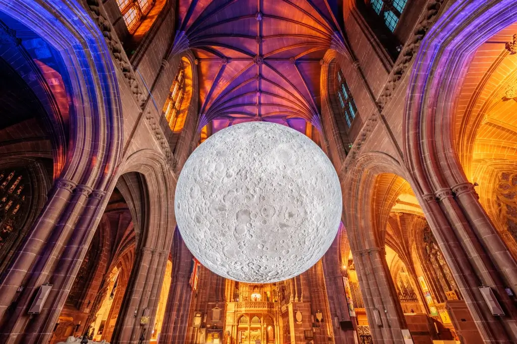 A large, realistic moon installation hangs inside a gothic cathedral, illuminated by colorful lights from the stained glass windows.