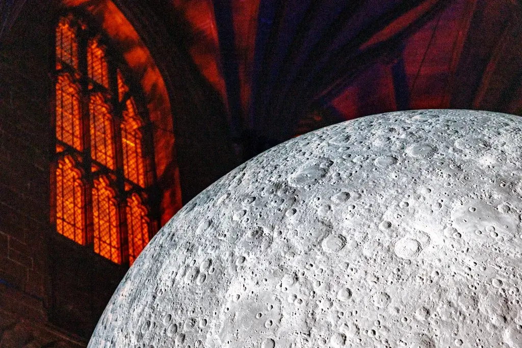A large, realistic moon installation hangs inside a gothic cathedral, illuminated by colorful lights from the stained glass windows.