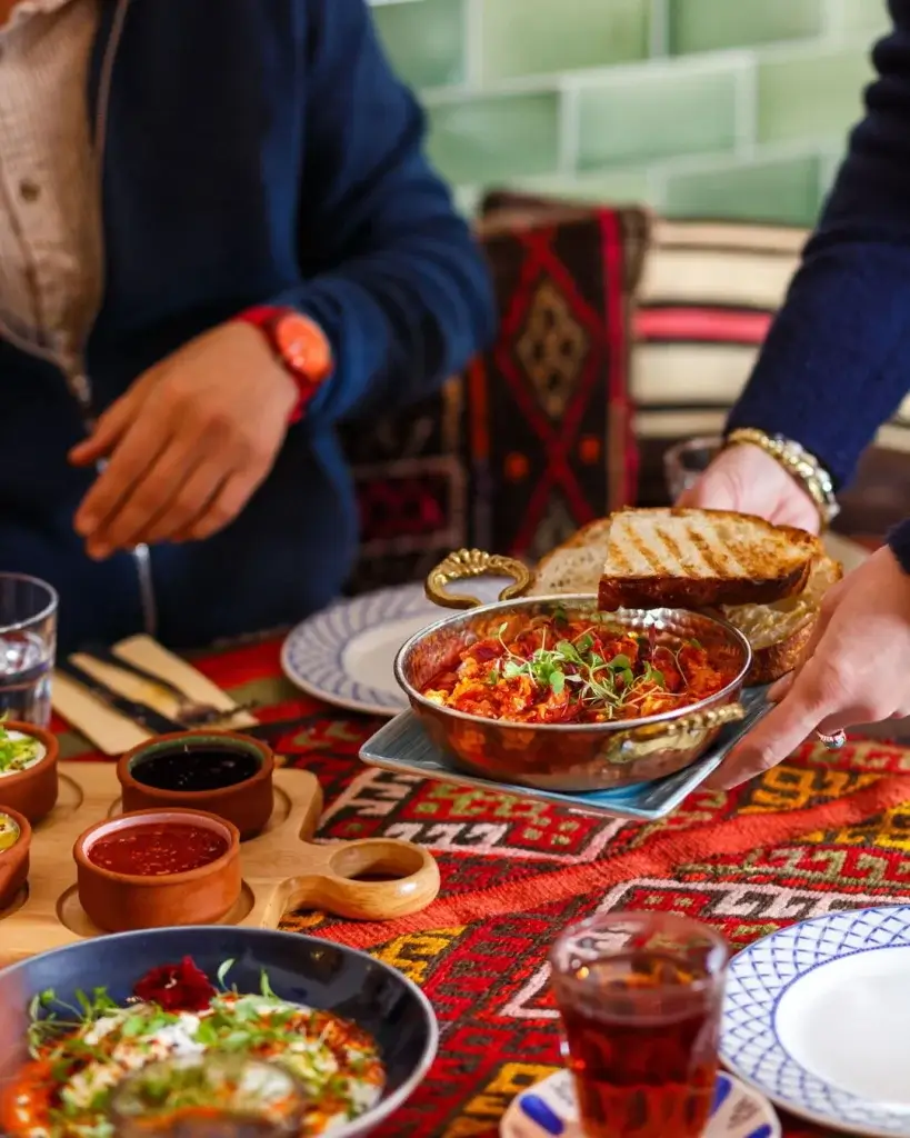 Hands serving a copper pan of tomato stew with microgreens and grilled bread on a colourful patterned table beside sauces and tea.