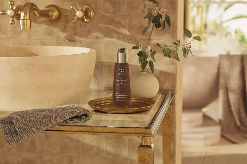 A bottle of massage body oil by Rituals is displayed on a gold tray beside a textured towel and a decorative plant in a spa-like setting.