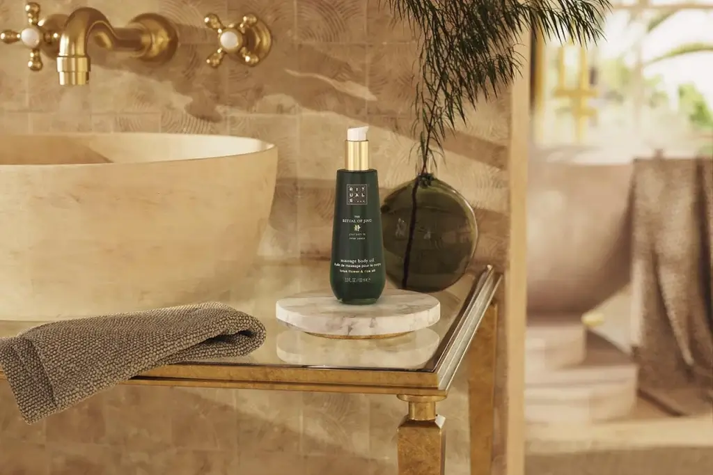 A bottle of massage body oil by Rituals is displayed on a marble tray beside a textured towel and a decorative plant in a spa-like setting.