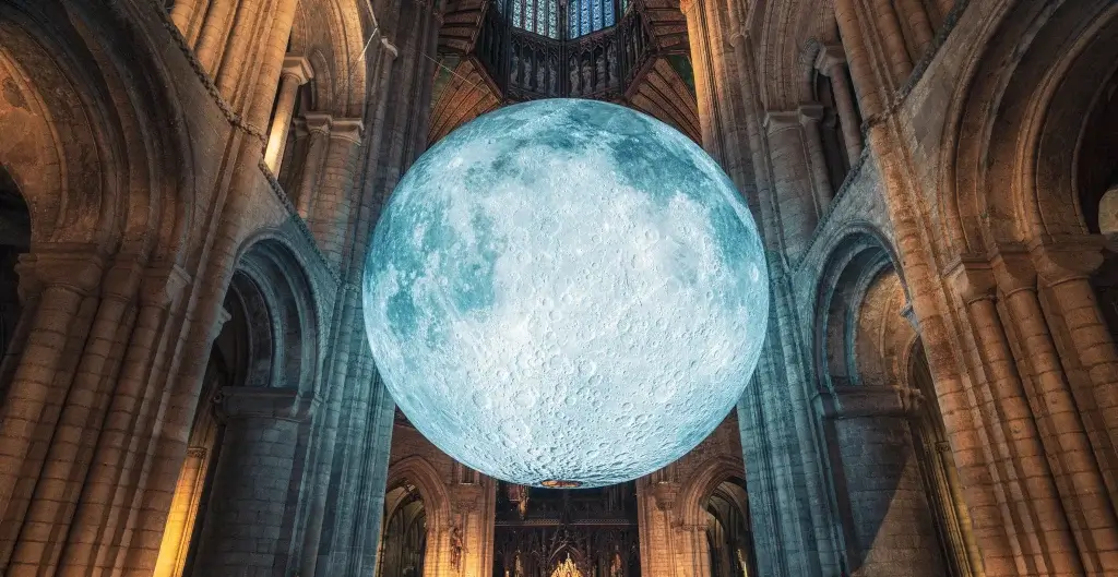 A large, illuminated moon model hangs in a grand cathedral, surrounded by stone arches and intricate details, creating an ethereal atmosphere.