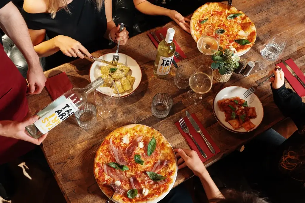 Group sharing an Italian meal: two pizzas and pasta on a wooden table, hands serving and white wine being poured.