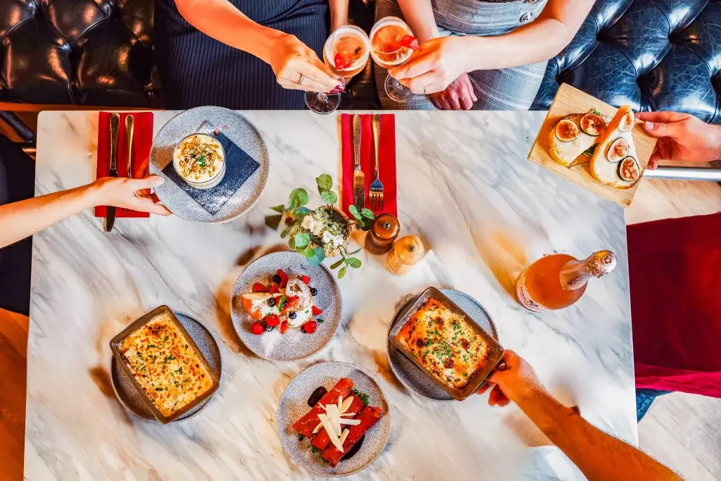 Overhead view of friends clinking cocktails over a marble table set with baked gratins, bruschetta and desserts.