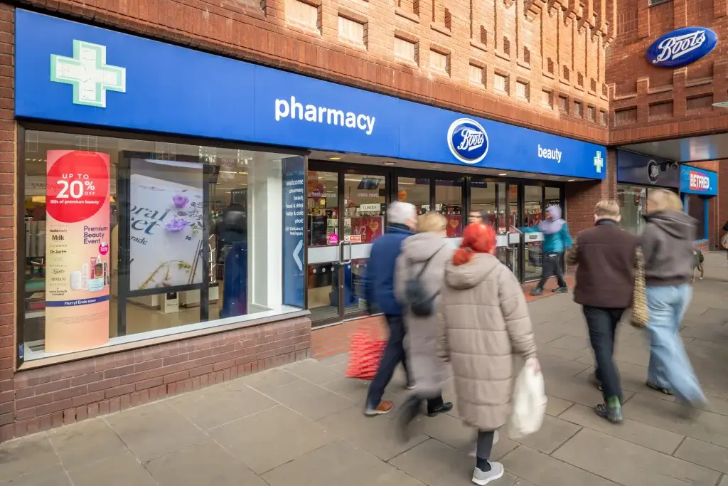 Boots pharmacy storefront with blue signage reading pharmacy and beauty, shoppers walking past on the pavement.