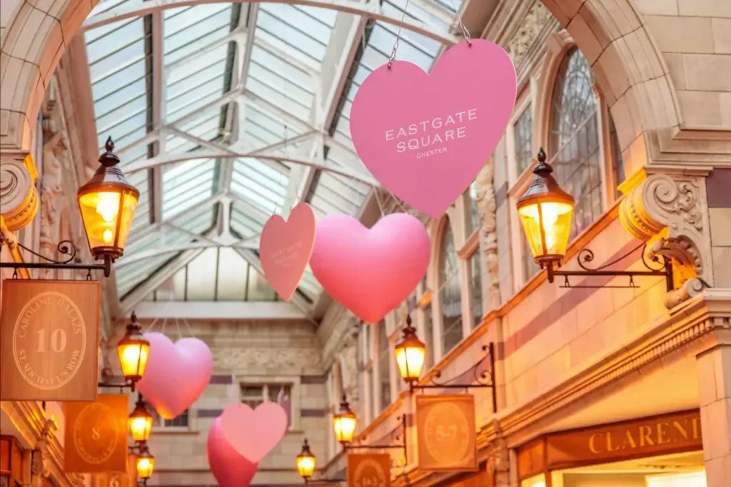 Large pink heart-shaped signs reading Eastgate Square suspended in a glass-roofed shopping arcade lit by warm lanterns.