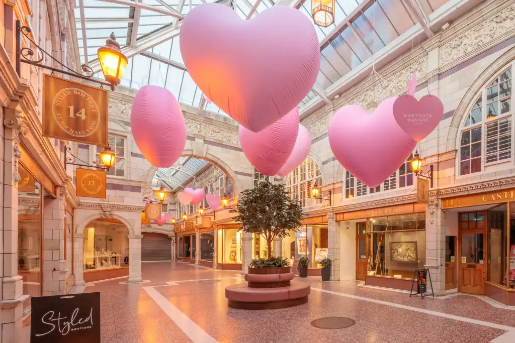 Victorian-style arcade with oversized pink heart balloons hanging from a glass roof over a central tree and seating.