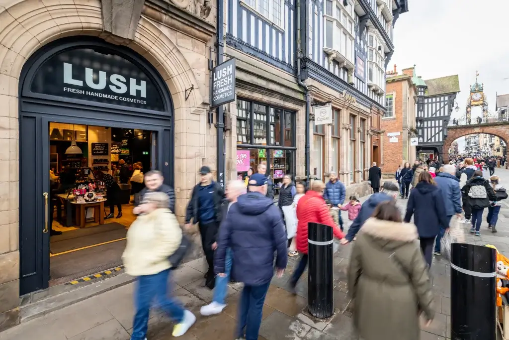 LUSH cosmetics shop with arched entrance on a busy historic high street, shoppers passing under a clocked arch in background.
