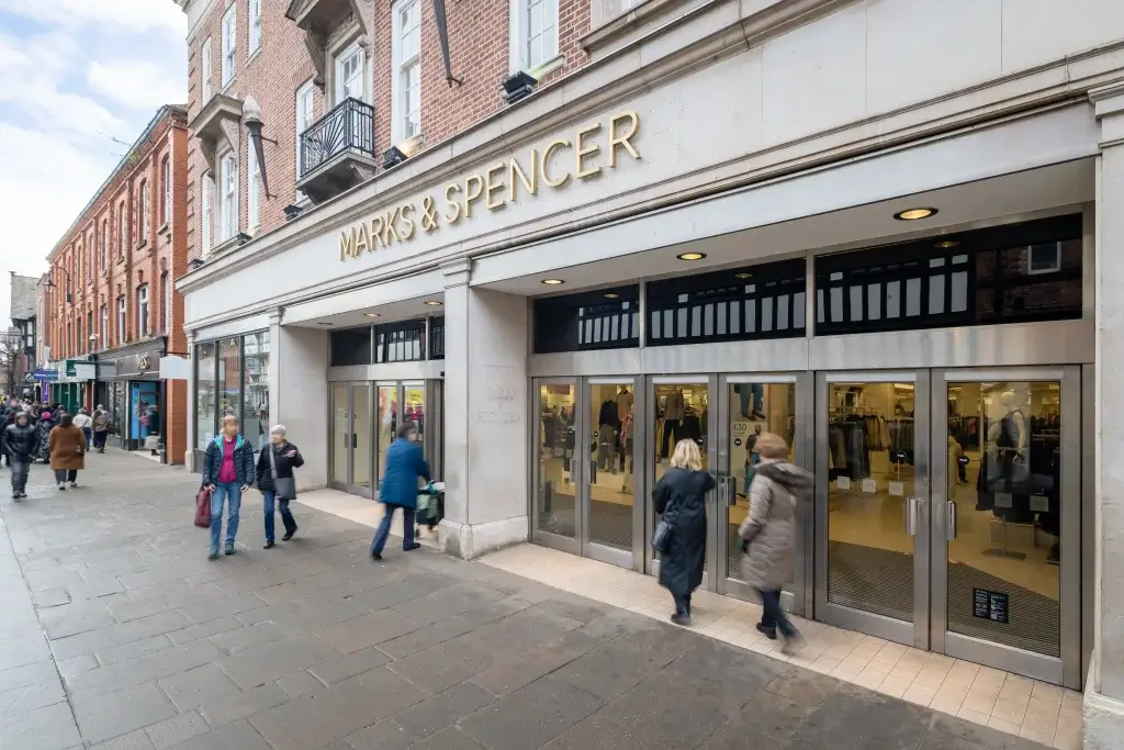 Marks & Spencer storefront with gold lettering above glass doors on a busy high street as shoppers enter.