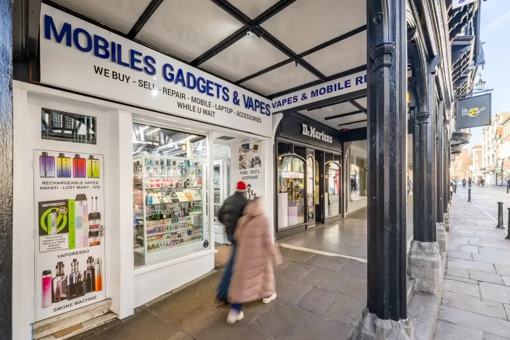 Shopfront of Mobiles Gadgets & Vapes with blue sign, vape and phone displays visible and blurred pedestrians walking past.