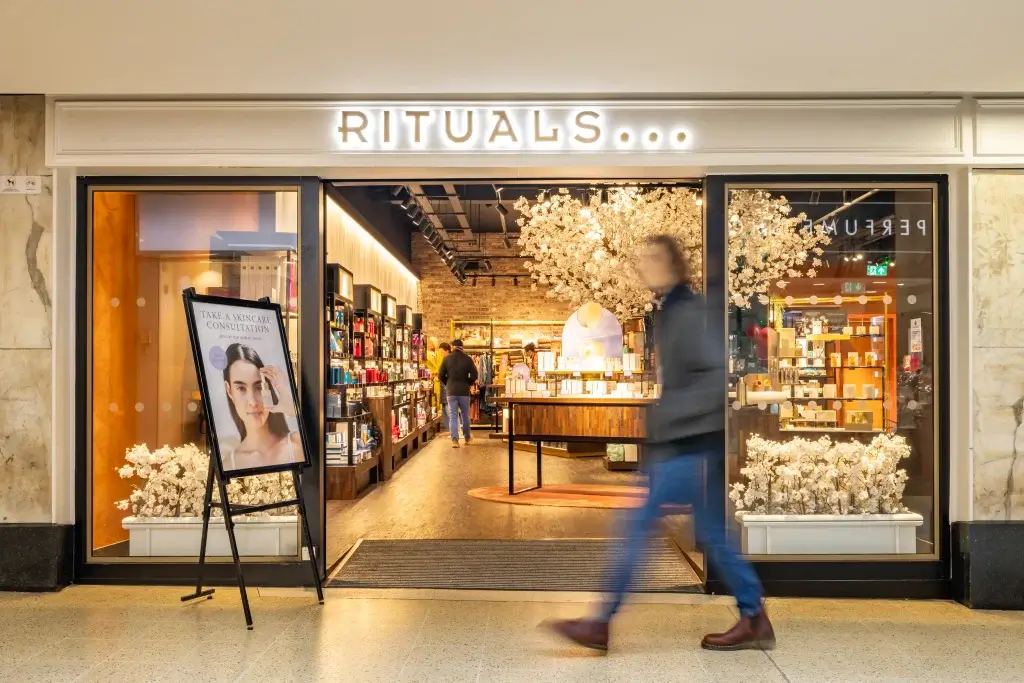 Rituals store entrance with illuminated sign, interior product shelves and blossom displays, skincare poster, blurred passerby.