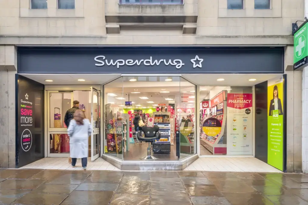 Superdrug storefront with black sign and star logo, glass doors revealing shelves and a salon chair as two people enter.