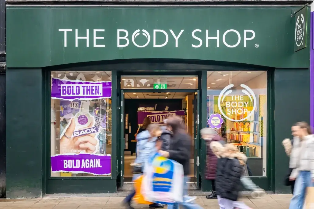 The Body Shop storefront with green sign, window posters reading Bold then Bold again and blurred shoppers passing by.