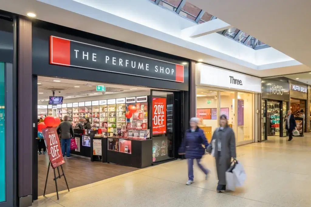 The Perfume Shop storefront in a mall with red 20% off sale signs and red balloons, shoppers walking past.