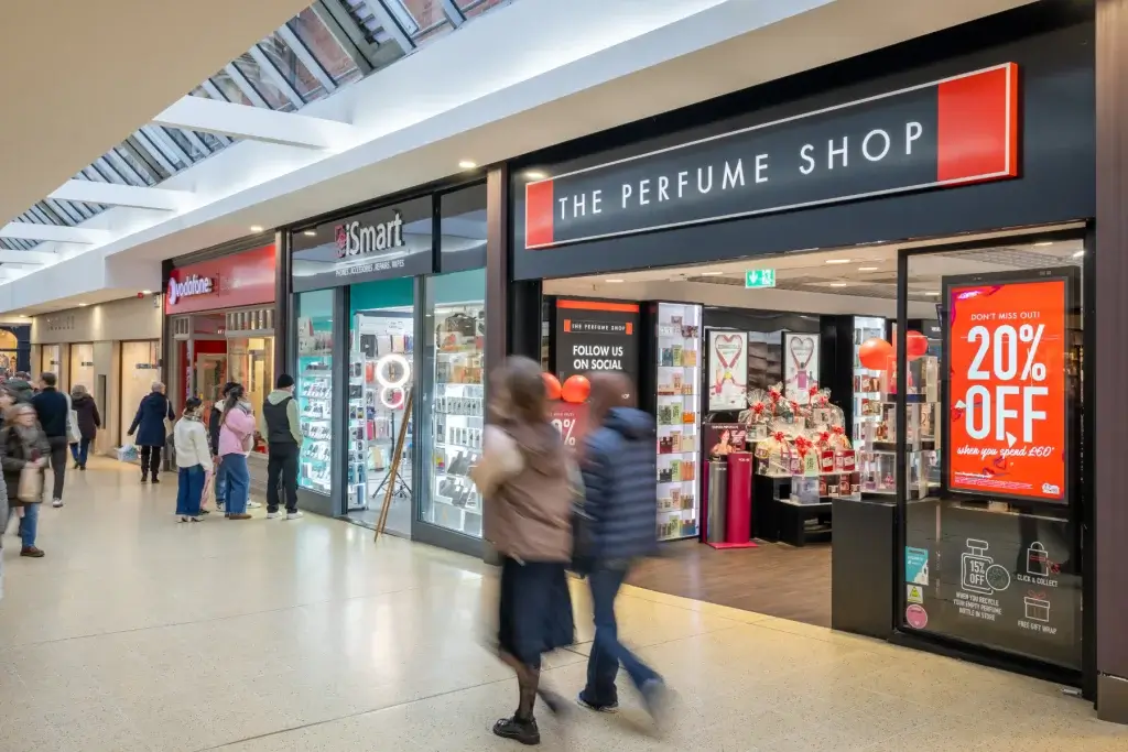 The Perfume Shop storefront in a shopping centre with a large red 20% OFF promotion display and shoppers walking by.