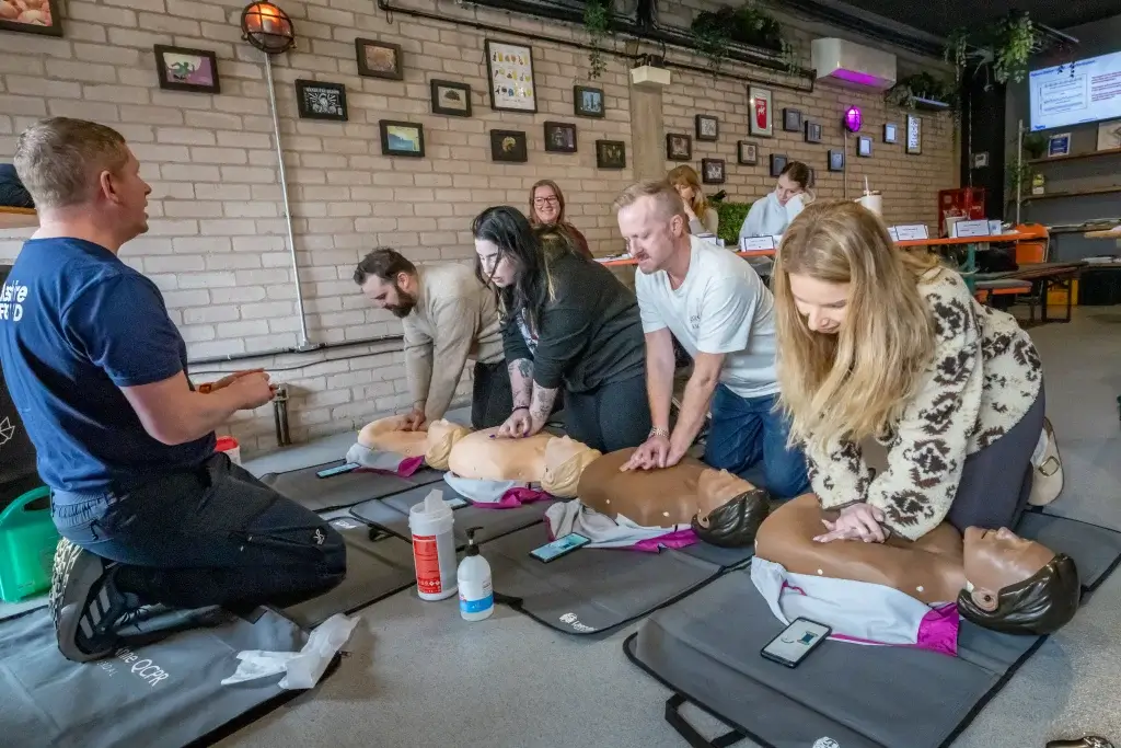 A group of people practicing CPR on mannequins during a training session in a room with framed pictures on the wall.