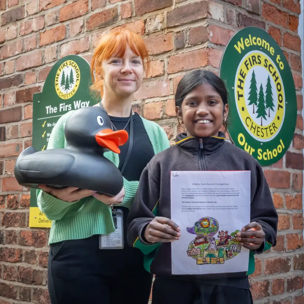 Two people stand in front of a brick wall with "The Firs School" signs, one holds a large rubber duck, the other a colorful duck drawing.