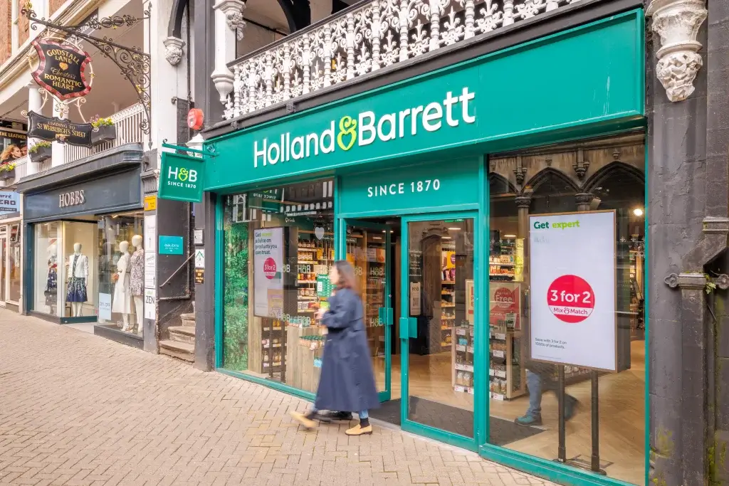 Exterior view of a Holland & Barrett health store with a woman walking past the entrance on a paved sidewalk.