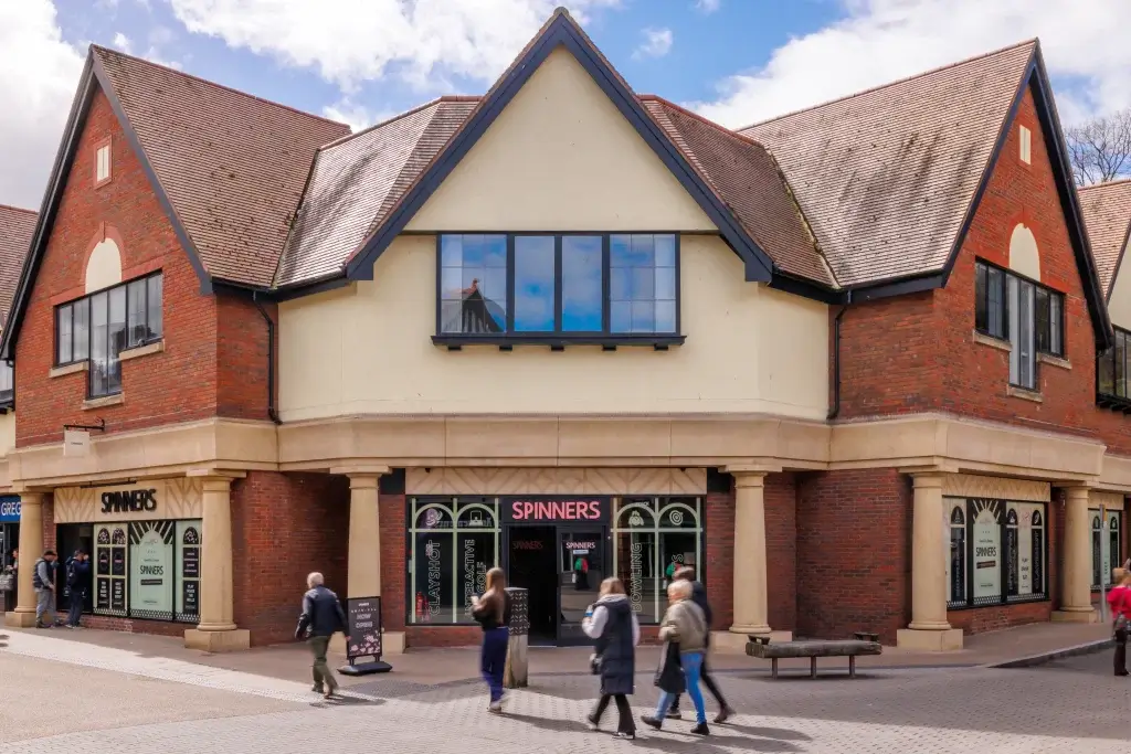 Two-storey red-brick building with peaked gables and central SPINNERS arcade entrance, shoppers walking by.