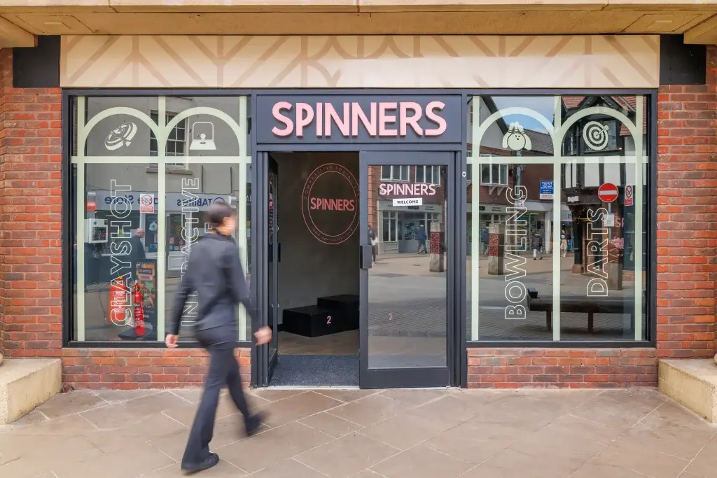 Spinners storefront with pink sign, glass windows listing bowling and darts, and a blurred pedestrian walking past.