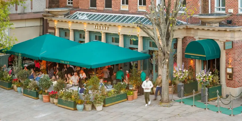 Outdoor dining terrace of The Ivy with large green canopies, potted plants and seated patrons.