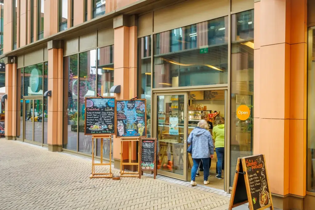 Cafe storefront with large glass windows, colourful chalkboard menus on easels and customers entering through sliding doors.
