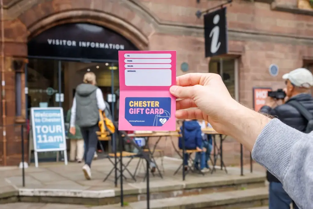 Hand holding a pink Chester gift card booklet in front of a visitor information centre entrance with people nearby