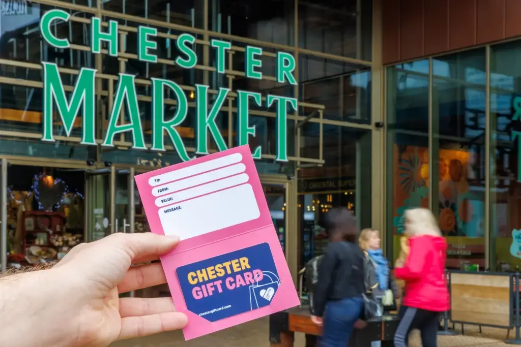 Hand holding a pink Chester gift card holder showing the card outside the green "Chester Market" entrance with blurred shoppers in the background.