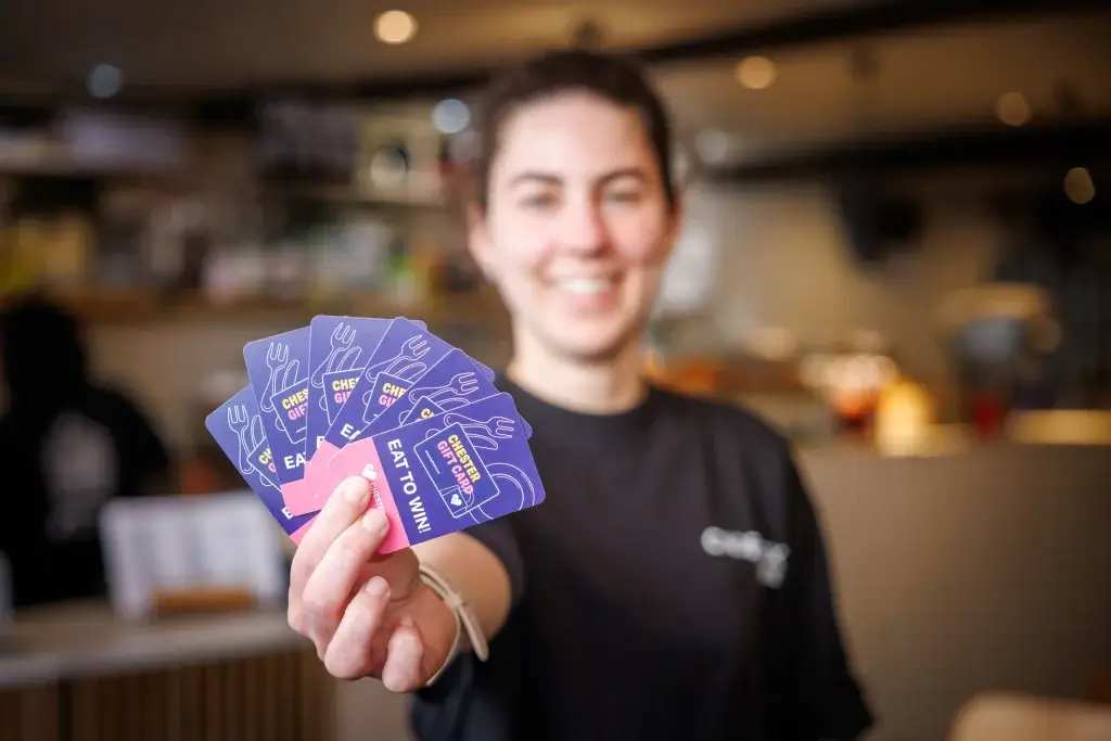 Smiling person in a café holding a fan of purple and pink gift cards that read "EAT TO WIN"