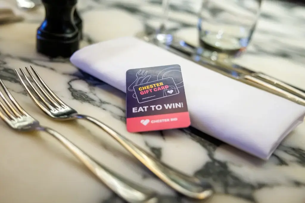 Chester gift card reading "EAT TO WIN!" on a folded white napkin atop a marble table, with forks and a glass nearby.