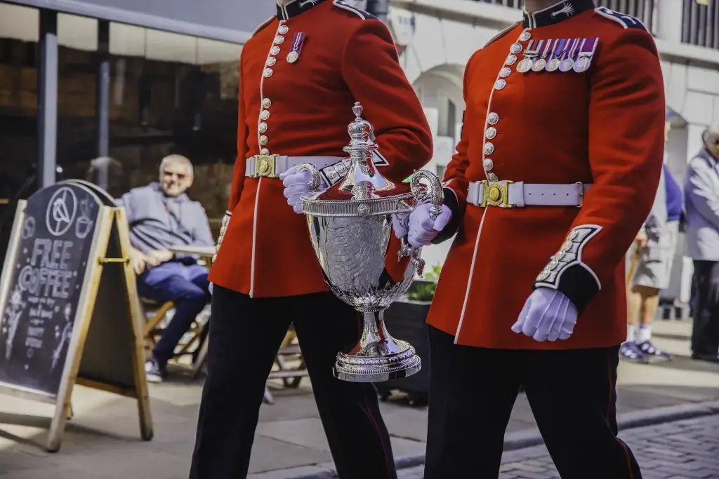Two soldiers in red ceremonial coats and white gloves carry a large ornate silver trophy, medals visible on their chests.