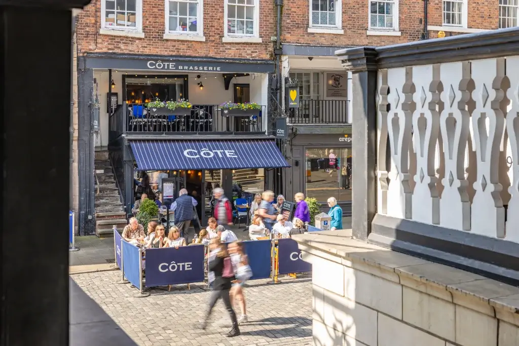 Outdoor seating at Côte Brasserie with blue striped awning, diners at tables and blurred pedestrians on a cobbled street.