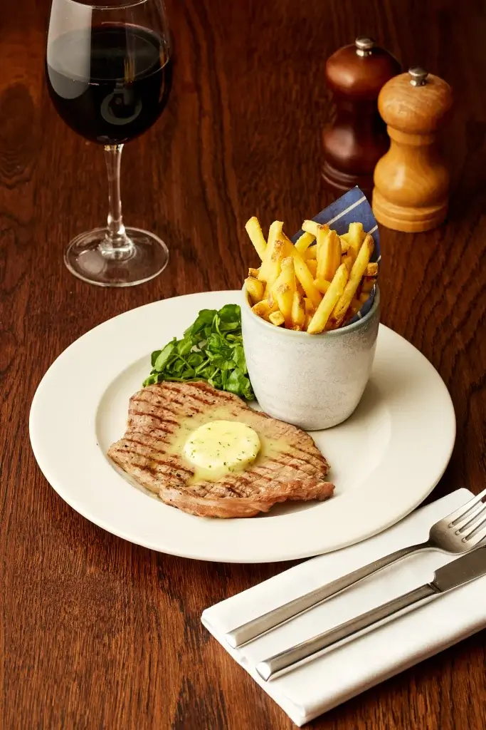 Grilled steak with melting herb butter, fries in a cup and watercress, with red wine and cutlery on a wooden table.