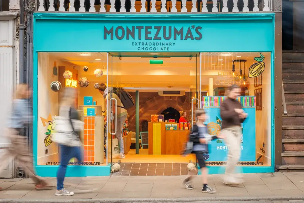 Turquoise Montezuma's chocolate shopfront with glass doors and blurred pedestrians passing by.