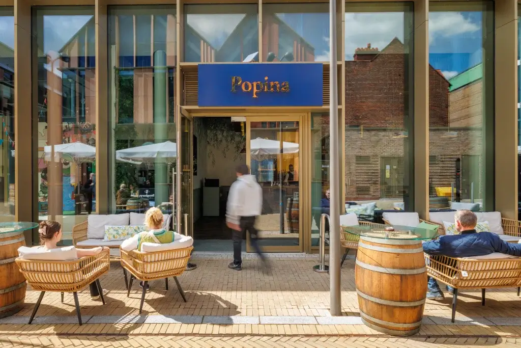 Cafe entrance with blue Popina sign above glass doors, outdoor wicker seating, barrel tables and patrons.