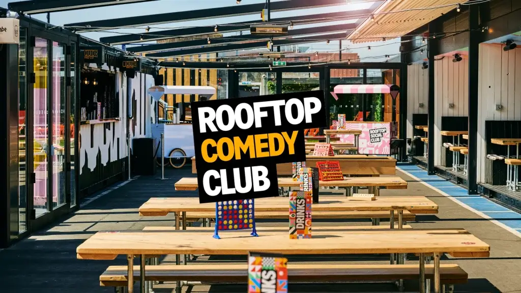 Large Rooftop Comedy Club sign above wooden picnic benches and colourful stalls under a pergola with string lights