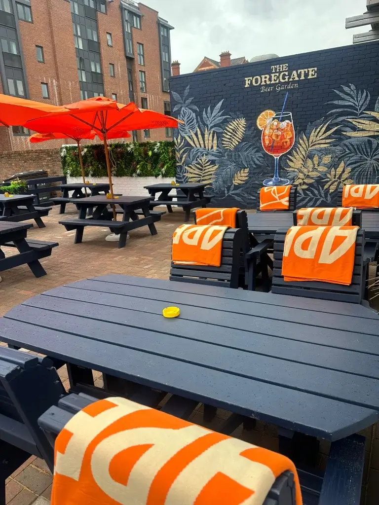 Beer garden with navy picnic tables, orange umbrellas and blankets, and a wall mural reading The Foregate with a cocktail.