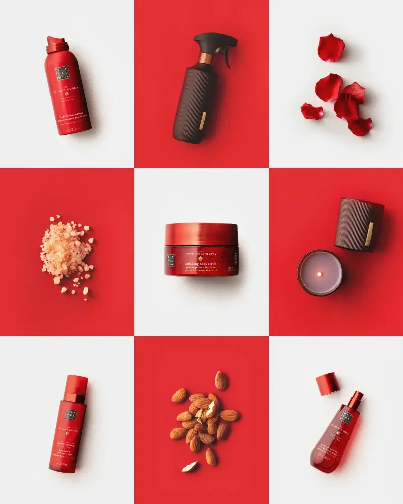 Flatlay of red skincare products, jar, sprays, candle and rose petals with almonds and salt on a red-and-white checkerboard.