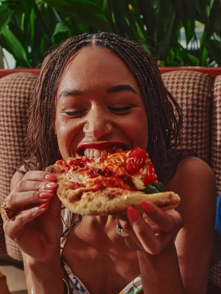 Young woman with braided hair and nose ring smiling with eyes closed as she bites a tomato-topped pizza slice.