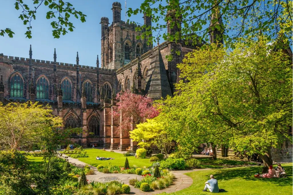 Gothic cathedral facade with tall arched windows framed by a sunny walled garden where people sit and relax on the lawn